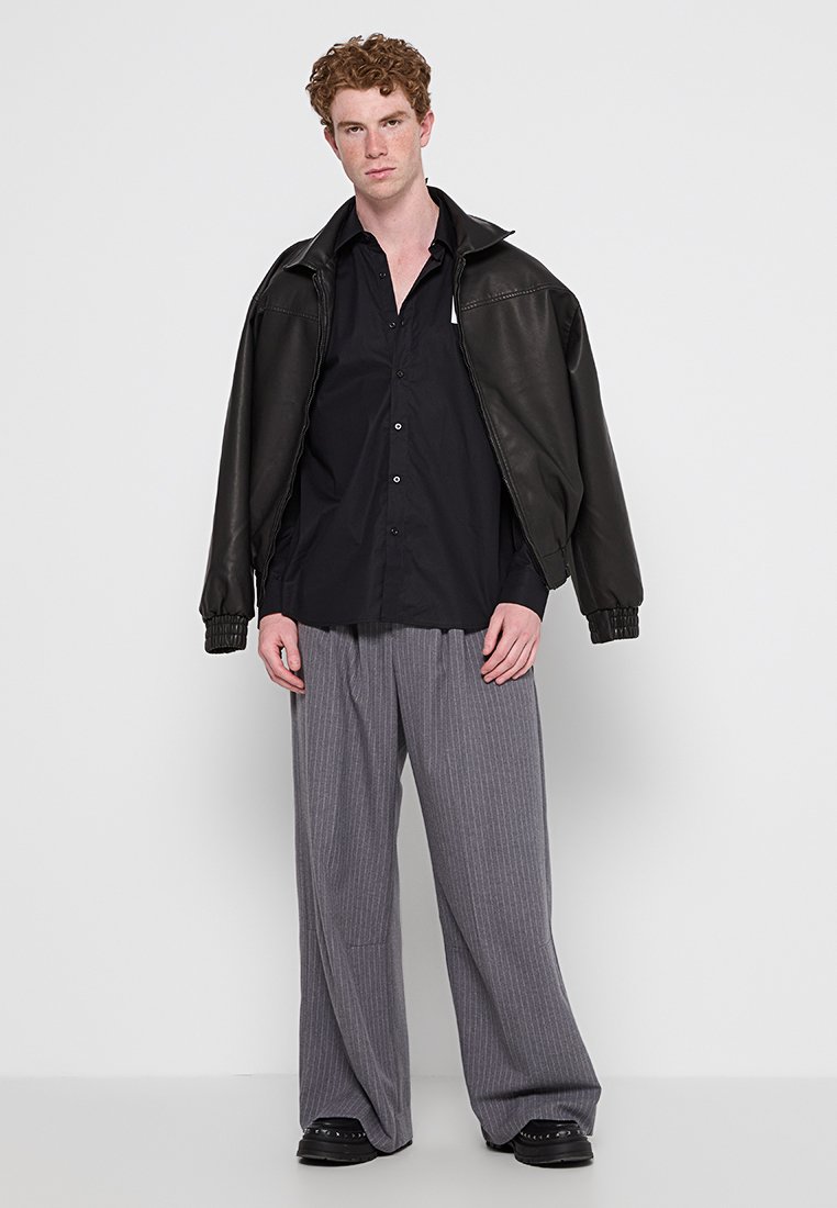 Veste bomber en cuir noir, chemise noire à boutons, pantalons larges gris à rayures, et chaussures à plateforme noires. Design simple et structuré.