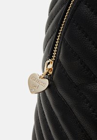 Čierna prešívaná kabelka s výrazným zlatým zipsom a srdiečkovým príveskom označeným "GUESS Girl", ktorý má malý diamantový akcent.