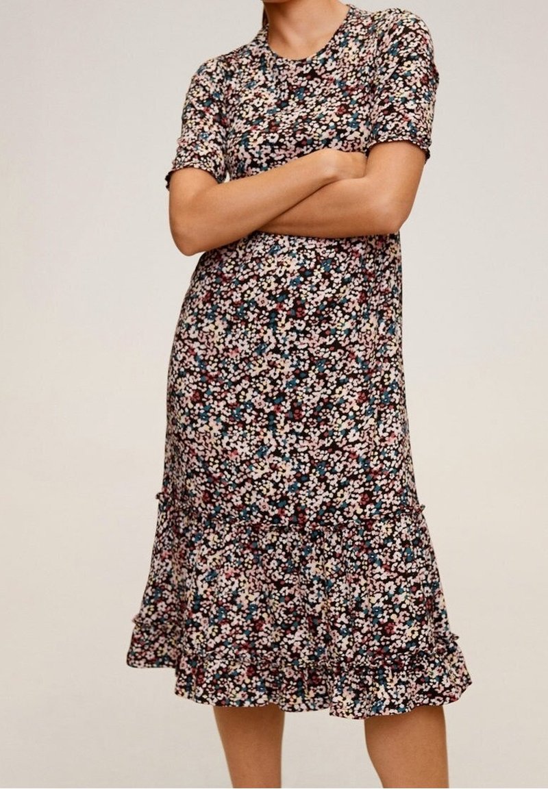 Femme portant une robe midi à manches courtes et imprimé floral avec un ourlet volanté, debout les bras croisés devant un fond uni.