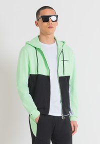 Felpa con zip mint green e nera con cappuccio, dotata di dettagli riflettenti e lacci, indossata sopra una maglietta bianca e pantaloni neri.