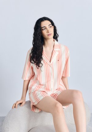 Jong persoon met lang krullend haar zit op een lichtgrijze stoel met textuur, draagt een lichtroze en witte gestreepte korte mouwen shirt en bijpassende korte broek.