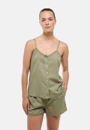 Femme portant un débardeur vert olive boutonné et un short assorti avec bordure en dentelle, debout avec la main dans la poche devant un fond uni.