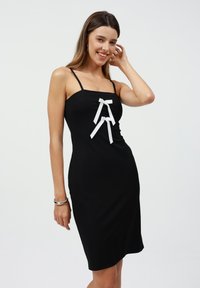 Vestito nero senza maniche con una vestibilità dritta, caratterizzato da due fiocchi bianchi come accenti sul davanti. Tessuto di consistenza liscia.