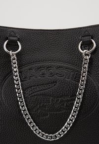 Sac en cuir noir texturé avec un logo en relief bien visible, une bandoulière chaîne argentée et des accents de ferrures métalliques.