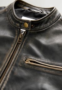 Zwarte leren jas met hoge kraag, bronzen hardware, een centrale rits en een borstzak met ritssluiting. Gelaagd oppervlak detail.