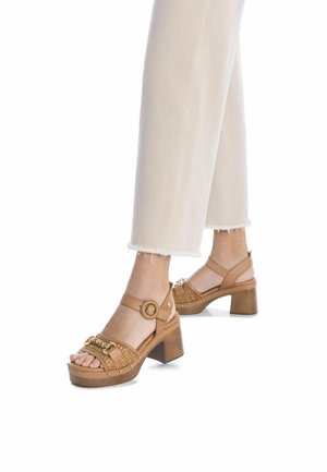Sandali con tacco beige con dettagli intrecciati, punta arrotondata, cinturino alla caviglia regolabile e tacco a blocco in legno chunky. Presentano piccoli accenti metallici.