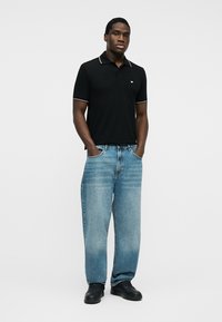 Άνδρας που στέκεται με τα χέρια στις τσέπες, φοράει μαύρο πόλο πουκάμισο, ανοιχτό μπλε τζιν και μαύρα παπούτσια σε απλό λευκό φόντο.