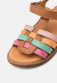 Sandal de cuero marrón con tiras tejidas multicolores en rosa, naranja y azul verdoso. Presenta una puntera redonda y una suela plana con detalles de costura.