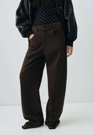 Pantalones anchos marrones de tela estructurada con dos bolsillos laterales, combinados con una camisa negra de manga larga con puntos y una chaqueta bomber negra.
