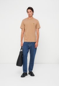 T-shirt beige à manches courtes avec un petit logo jaune, associé à un jean bleu et des chaussures noires, tenant un sac fourre-tout noir.