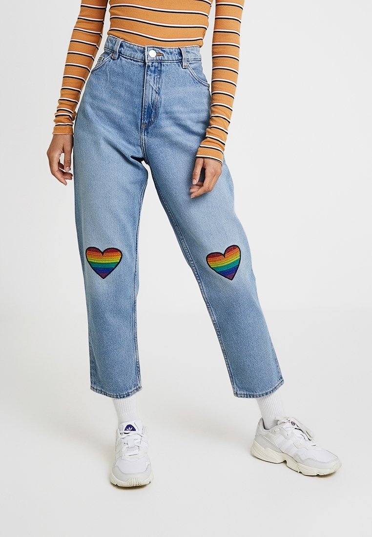 Personne portant un jean bleu clair avec des patchs cœur arc-en-ciel au niveau des genoux, associé à un haut marron rayé à manches longues et des baskets blanches.