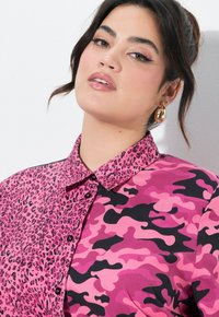 Rosa Hemd mit einem Leopardenmuster auf einer Seite und einem rosa Camouflage-Design auf der anderen. Verfügt über einen Kragen und Knöpfe.