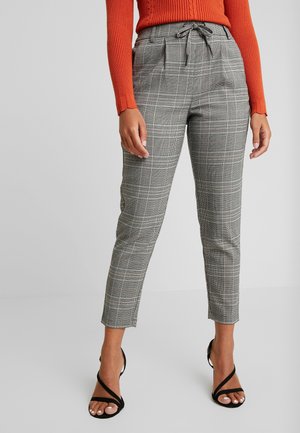 Femme portant un pantalon gris à carreaux fuselé avec une taille à cordon, un haut orange côtelé à manches longues, et des talons noirs à brides.