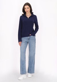Pullover lungo a maniche con colletto e mezza zip di colore blu navy, abbinato a jeans a gamba larga blu chiaro e sneakers bianche. Tessuto liscio e vestibilità casual.
