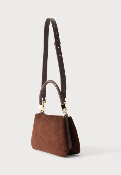 Sac d’épaule en daim marron avec une poignée courte tressée, une longue bandoulière réglable et des fermoirs dorés, présenté sur un fond uni.