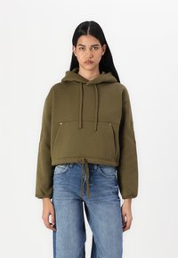 Ελαιότοξο κοντό φούτερ με κορδόνια, εμπρόσθιες τσέπες και κεντημένο λογότυπο στο μανίκι. Συνδυάζεται με ανοιχτό μπλε τζιν.