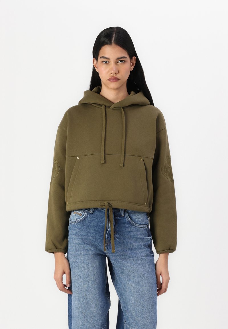 Ελαιότοξο κοντό φούτερ με κορδόνια, εμπρόσθιες τσέπες και κεντημένο λογότυπο στο μανίκι. Συνδυάζεται με ανοιχτό μπλε τζιν.