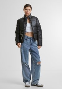 Kvinne i svart boblejakke, hvit crop top, revne vide jeans og hvite sneakers som står mot en ensfarget bakgrunn.