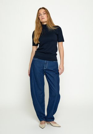 Žena s dugom kosom u tamnoplavoj majici kratkih rukava s visokim ovratnikom, širokim plavim trapericama i bež cipelama na petu sa špicastim vrhom, stoji ispred jednostavne svijetle pozadine.