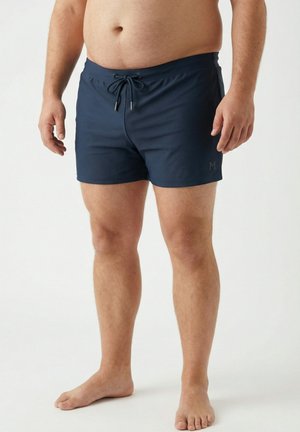 Uomo che indossa pantaloncini da bagno blu navy con coulisse, in piedi a piedi nudi su uno sfondo chiaro e uniforme.