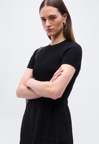 Γυναίκα με μακριά καστανά μαλλιά, φορώντας μαύρο κοντομάνικο τοπ και μαύρη υφασμάτινη φούστα, με σταυρωμένα χέρια, ασημένια σκουλαρίκια κρίκους και μαύρη τσάντα ώμου.