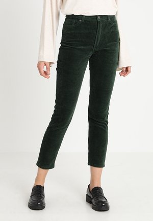 Vrouw die donker groene smalvallende corduroy enkellange broek draagt, een creme kleurige top met lange mouwen en zwarte loafers, staand tegen een witte achtergrond.
