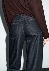 Person trägt dunkelblaue weit geschnittene Jeans und einen in die Hose gesteckten braunen Langarmpullover, steht vor einem hellen Wand Hintergrund.