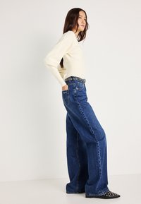 Κρεμ ζιβάγκο με μακριά μανίκια; ψηλόμεσες, φαρδιές μπλε τζιν παντελόνες; μαύρη ζώνη με κυκλική αγκύλη; μαύρα παπούτσια με μυτερή μύτη.