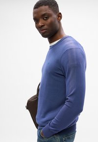 Blauer Rundhalsausschnitt-Pullover aus weichem Strick mit langen Ärmeln. Dezentes Logo-Detaill auf der Brust. Das Model trägt Jeans und trägt eine braune Tasche.