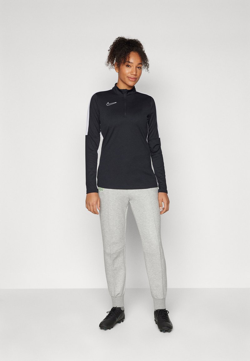 Donna in maglietta sportiva nera a maniche lunghe, pantaloni jogger grigi chiari e scarpe da ginnastica nere, in piedi contro uno sfondo bianco semplice.