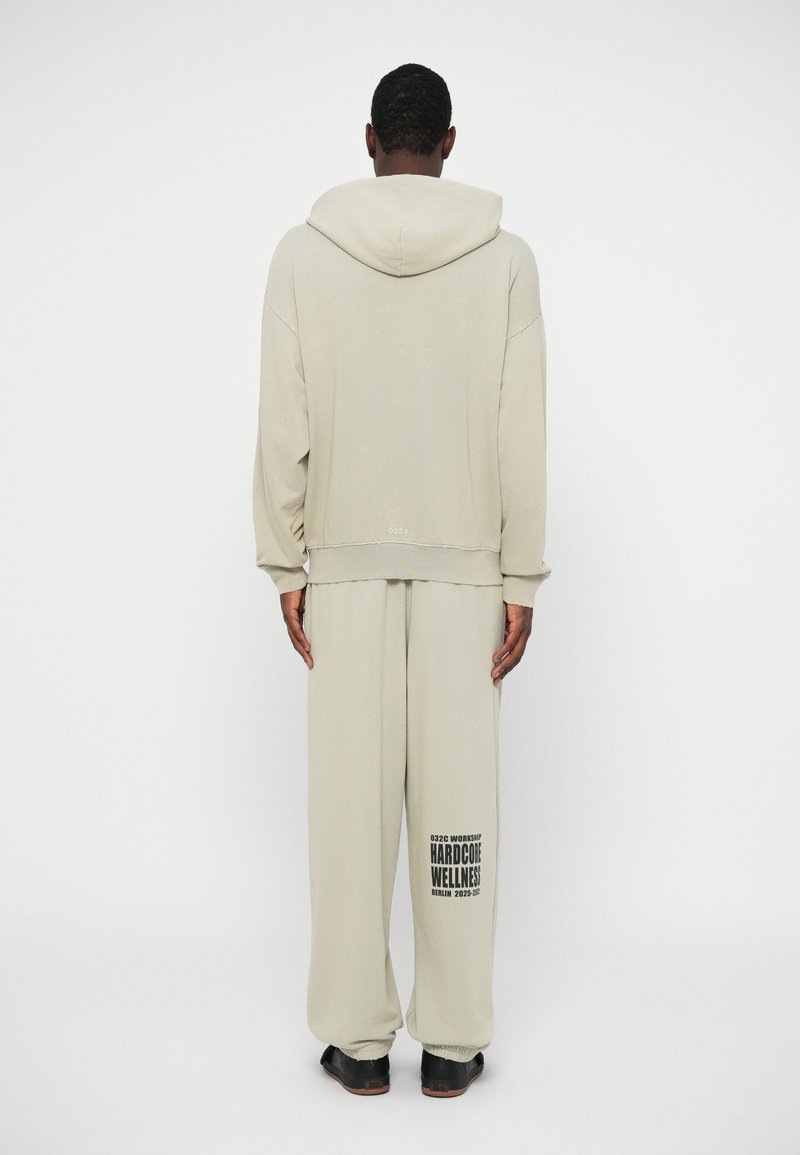 Persoon in lichtbeige hoodie en bijpassende sweatpants met de tekst "HARDCORE WELLNESS" op het linkerbeen, staand tegen een effen witte achtergrond.