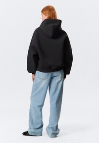 Črna oversized kapuca z nastavljivo kapuco, ki se ponaša s gladko teksturo in spuštenimi rameni, v kombinaciji s svetlomodrimi baggy jeans hlačami.