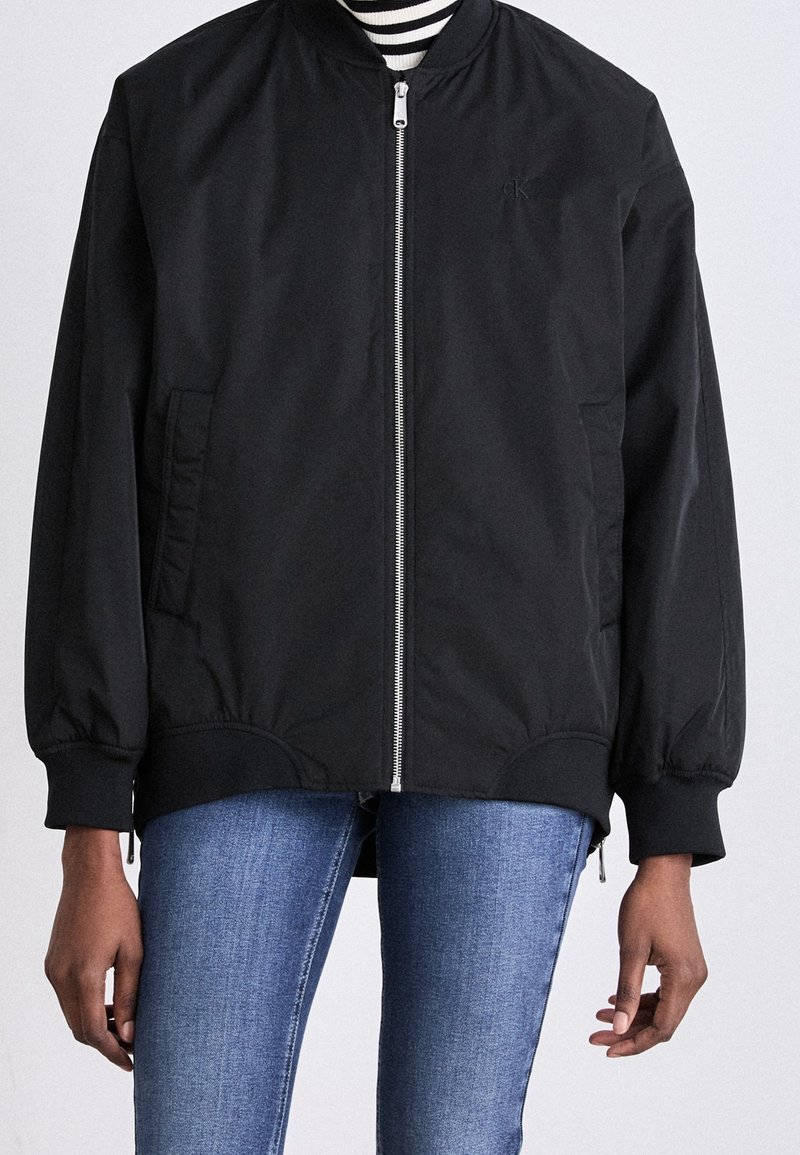 Cazadora bomber negra con cremallera, puños y bajo acanalados, llevada sobre una camiseta de rayas blancas y negras y combinada con jeans de mezclilla azul.