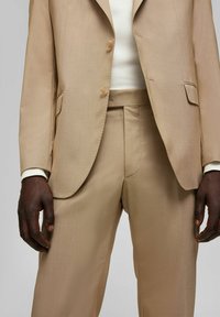 Beige op maat gemaakt pak van een gestructureerde stof, met een enkele knoopsluiting, scherpe revers en rechte broekspijpen. Witte shirt zichtbaar eronder.