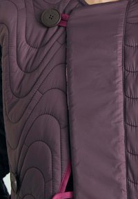 Fioletowa pikowana kurtka z przeszyciami w kształcie fal, dużymi ciemnymi guzikami oraz szerokim przednim panelem zakrywającym część klatki piersiowej.