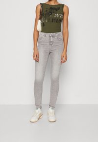 Top sin mangas de canalé verde oliva con estampado gráfico, combinado con jeans ajustados gris claro. Zapatillas blancas con acentos en beige completan el look.