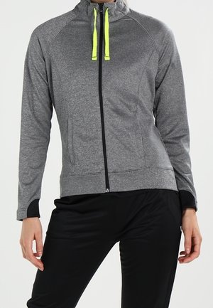 Person trägt eine graue Sportjacke mit Reißverschluss und neongelben Kordeln sowie schwarze Sporthose vor neutralem Hintergrund.