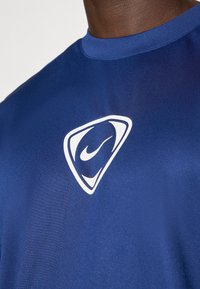 Μπλε αθλητικό μπλουζάκι κοντομάνικο, φτιαγμένο από λείο ύφασμα, με άσπρο λογότυπο σε τριγωνικό σχήμα στην περιοχή του στήθους.