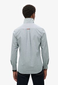 Lichtgrijs knopsluitend shirt met een hoge kraag en een lusdetail aan de achterkant. Voorzien van lange mouwen met knoopmanchetten en een gladde textuur.