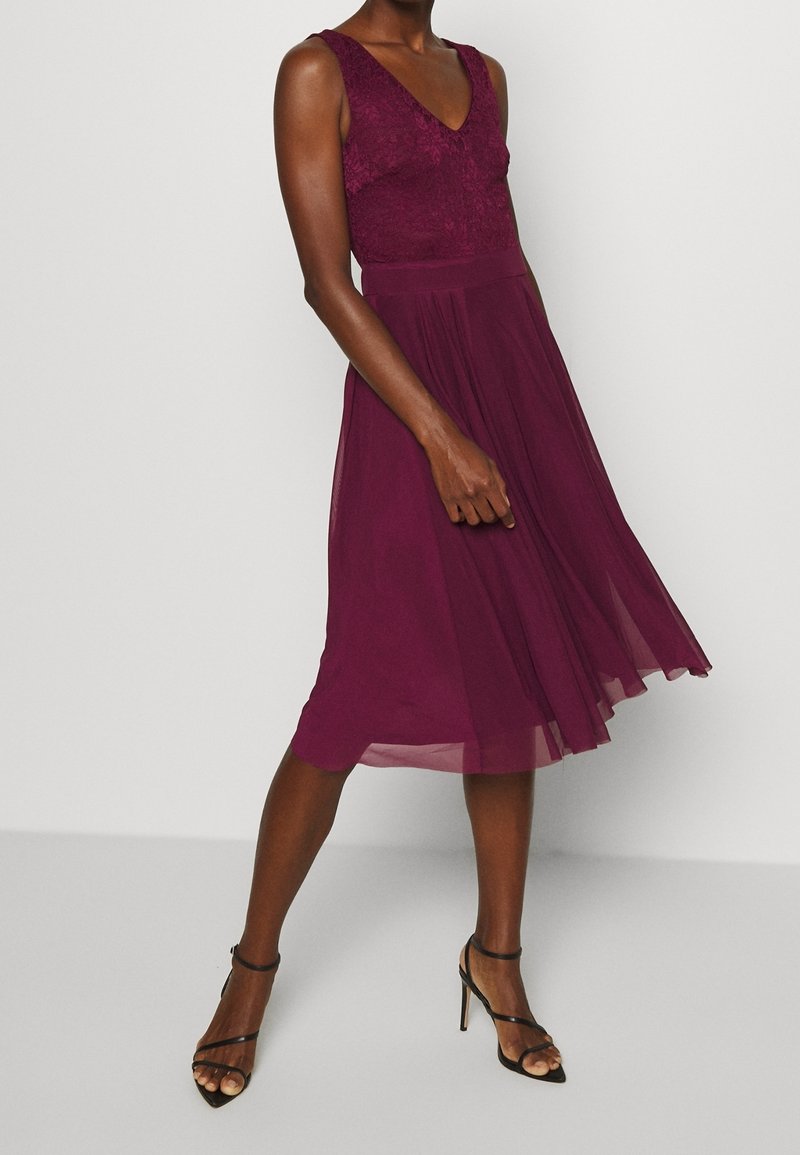 Femme portant une robe midi sans manches en dentelle et chiffon violet profond, avec des sandales noires à talons hauts à brides, debout devant un fond blanc.