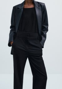 Zwarte leren blazer gecombineerd met een zwarte top en op maat gemaakte broeken. De blazer heeft een gestructureerd ontwerp en een soepele textuur; de broeken hebben een brede tailleband.