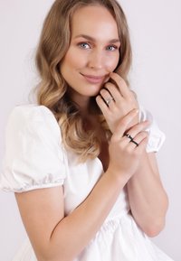 Zilveren en zwarte ringen gedragen op vingers met een gestructureerde afwerking, tegen een witte achtergrond. Het model heeft golvend haar en een lichte jurk.