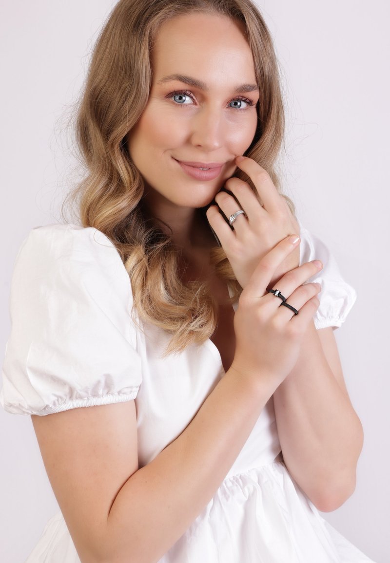 Zilveren en zwarte ringen gedragen op vingers met een gestructureerde afwerking, tegen een witte achtergrond. Het model heeft golvend haar en een lichte jurk.
