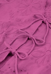 Roze stof met bloemenborduurwerk en decoratieve strikjes. De textuur lijkt zacht, met een gladde afwerking en geborduurde bladeren- en bloemenpatronen.