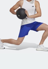 Person i vit linne och blå shorts som gör ett utfall medan de håller en svart Adidas medicinboll, iklädd vita träningsskor.