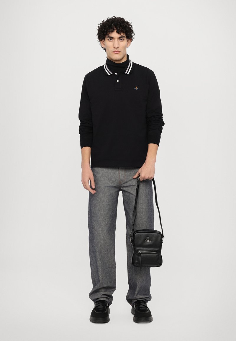 Zwarte polo met lange mouwen en witte strepen, grijze rechte jeans, zwarte tas en zwarte schoenen. Model poseert op een effen achtergrond.
