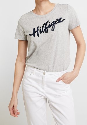 Jasnoszary t-shirt z krótkim rękawem z napisem "Hilfiger" w ciemnym stylu pisma, zestawiony z wysokim stanem białych spodni, jedna ręka w kieszeni.