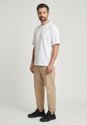 Skægget mand, stående iført en hvid t-shirt med korte ærmer, beige bukser med opsmøgede bukseben og sorte sneakers mod en ensfarvet lys baggrund.