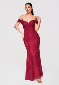 Femme dans une robe en dentelle bordeaux ajustée, épaules dénudées, corsage froncé, debout avec une main sur la hanche et l'autre près du visage.