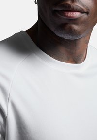 Biały krótki rękaw koszuli z teksturowanej tkaniny. Posiada okrągły dekolt i rękawy raglanowe. Widoczne tatuaże w okolicy szyi.